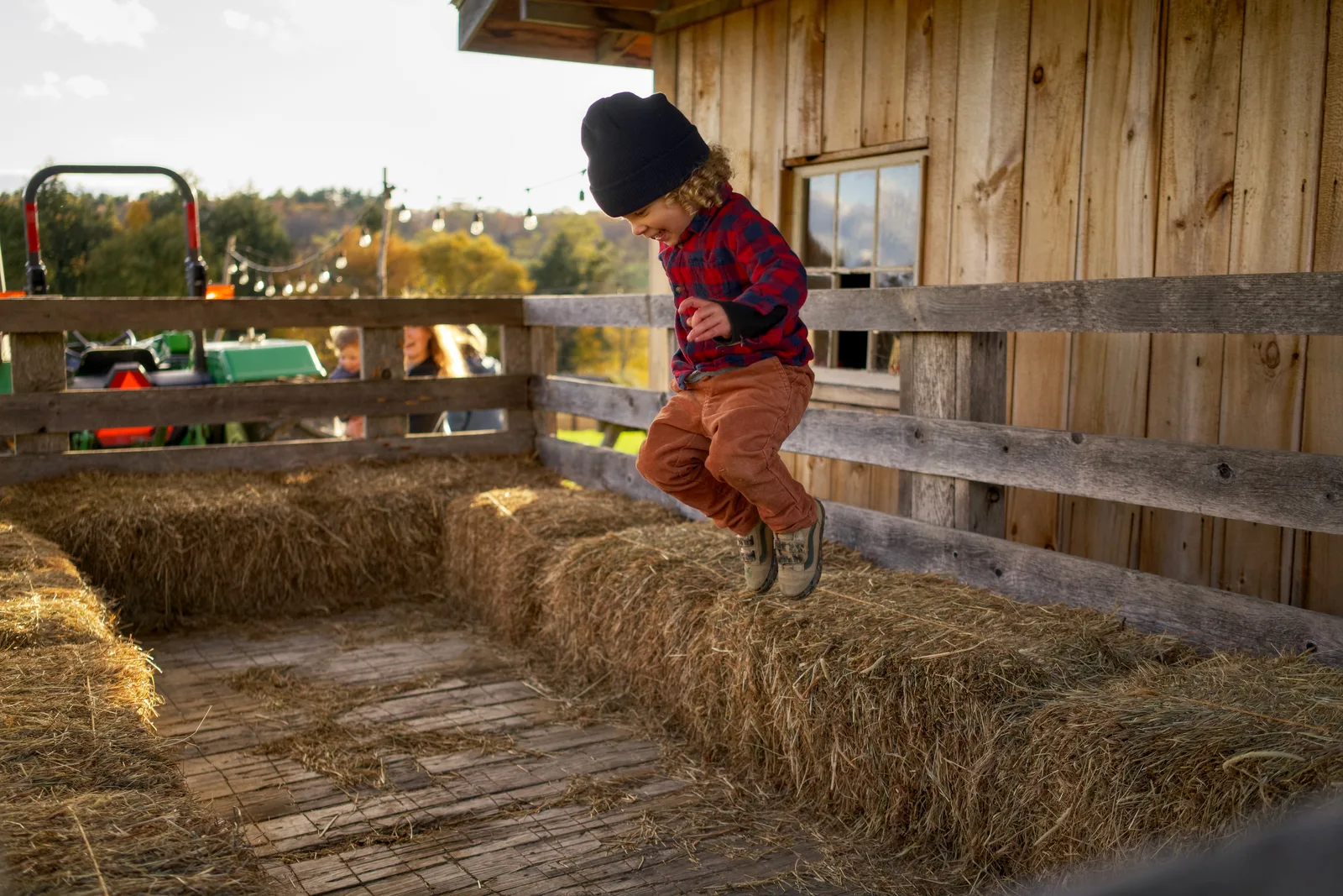 Child in black beanie, red plaid shirt, and orange pants landing on hay bales