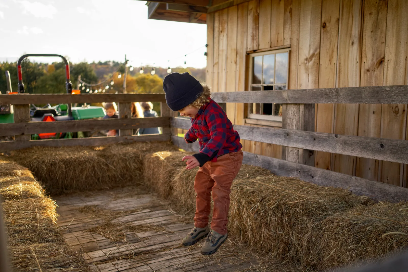 Child in black beanie, red plaid shirt, and orange pants bending over hay bales