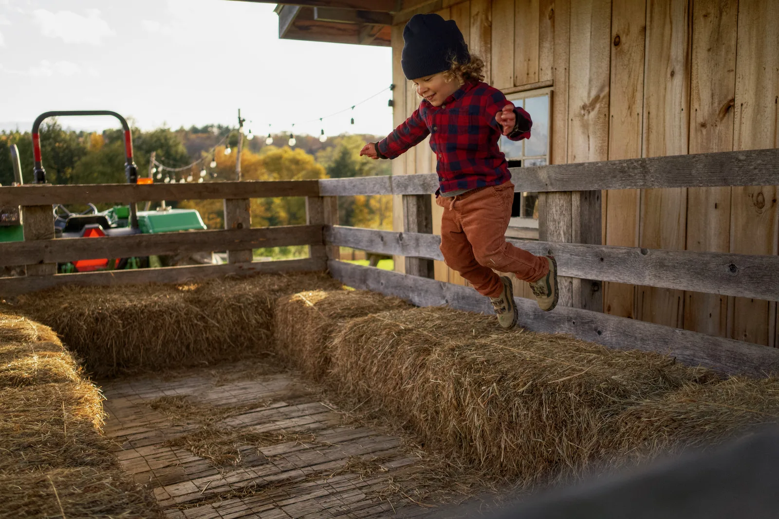 Child in black beanie, red plaid shirt, and orange pants leaping from hay bales