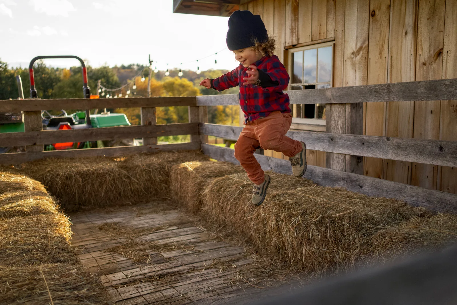 Child in black beanie, red plaid shirt, and orange pants jumping off hay bales