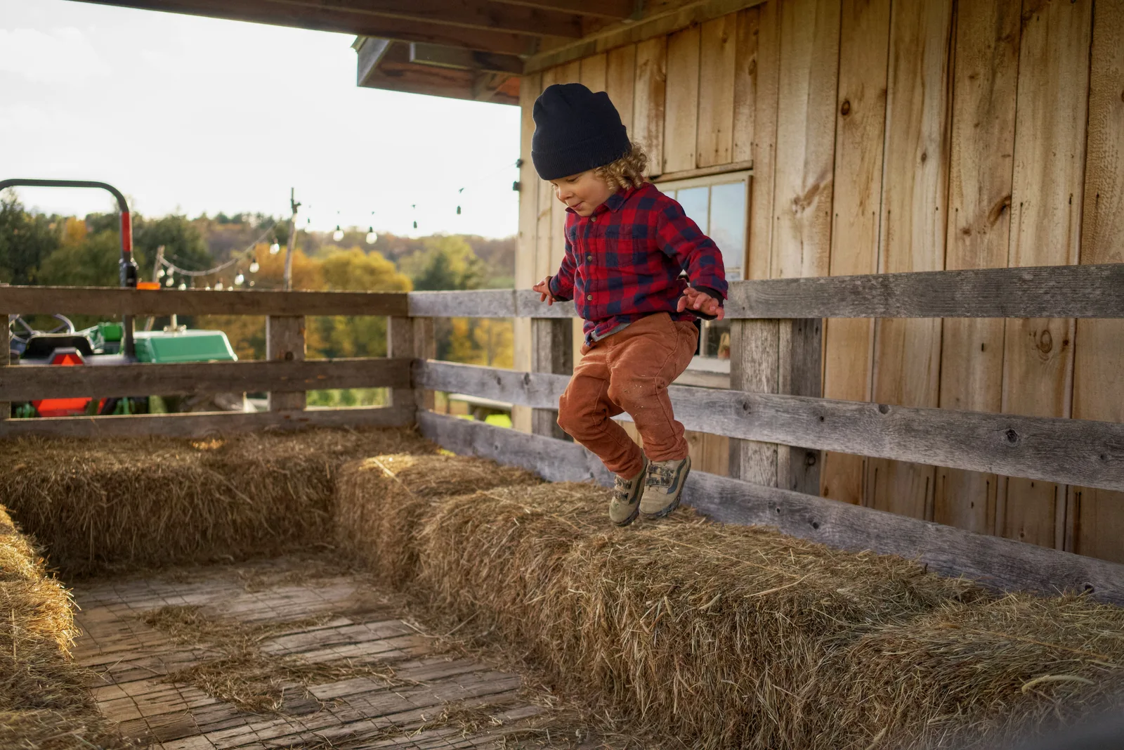 Child in black beanie, red plaid shirt, and orange pants stepping on hay bales