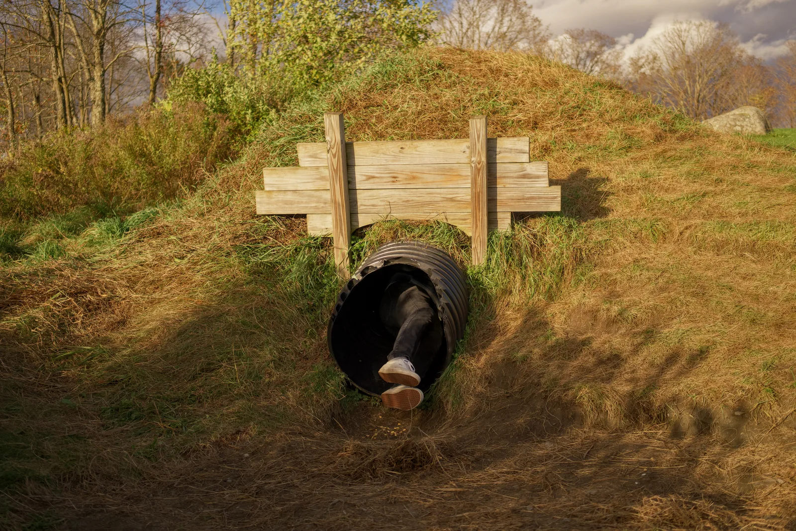 Person crawling through black culvert under wooden fence in grassy field
