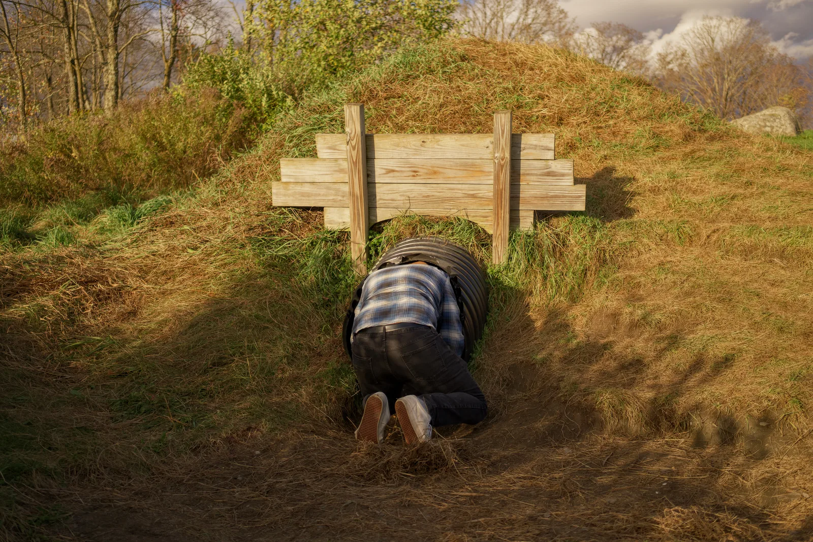 Person crawling through black culvert under wooden fence in grassy field