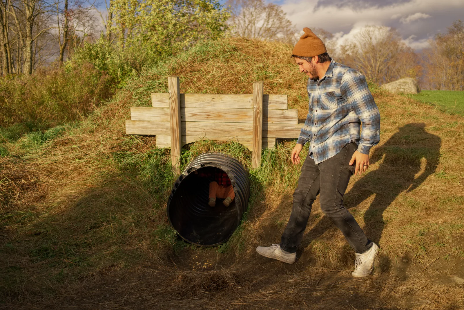 Adult in plaid shirt and brown beanie walking past culvert in grassy area