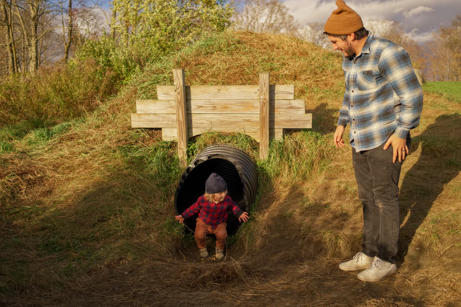 Child in red plaid shirt and brown pants emerging from culvert, adult nearby