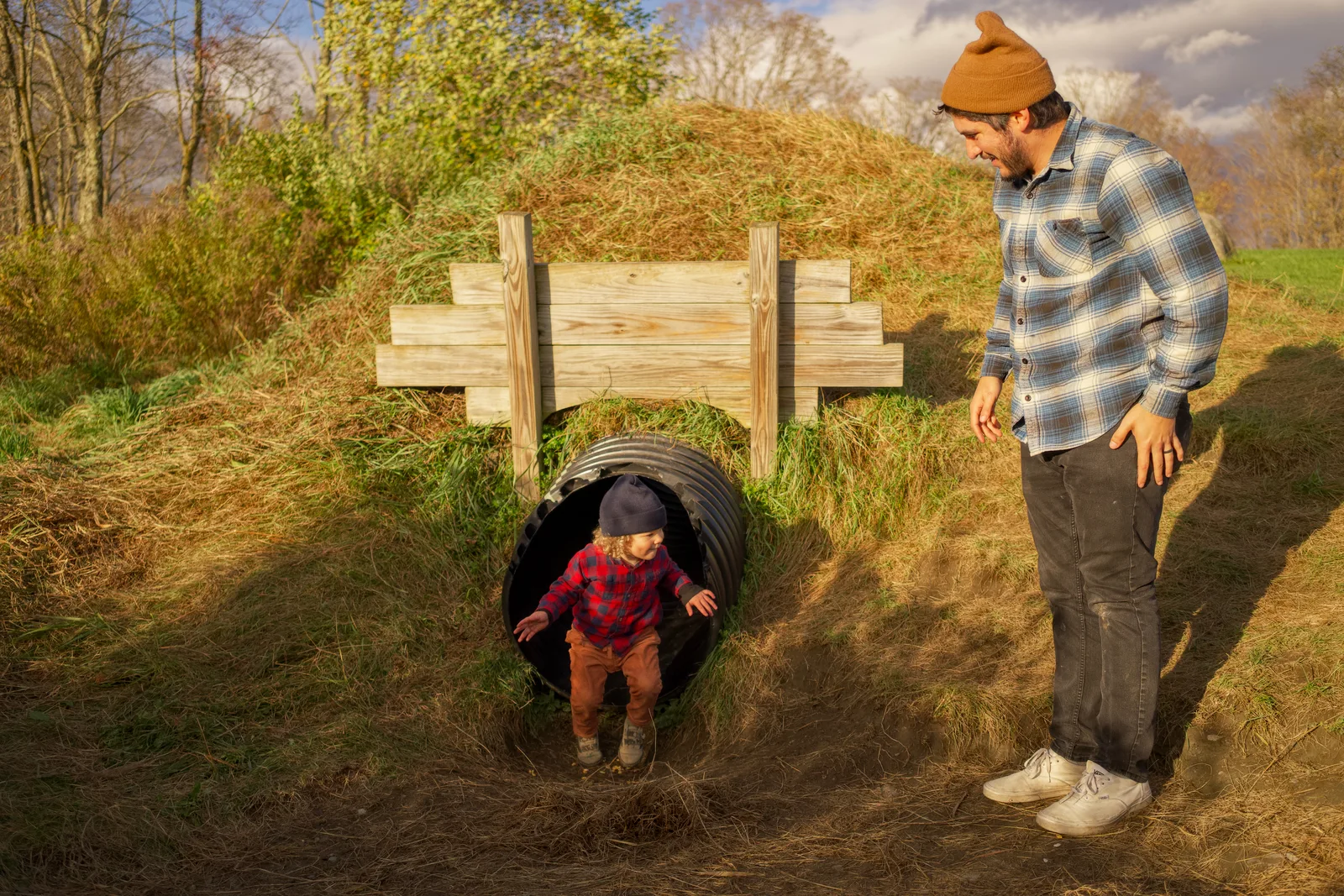 Child in red plaid shirt and brown pants exiting culvert, adult standing beside