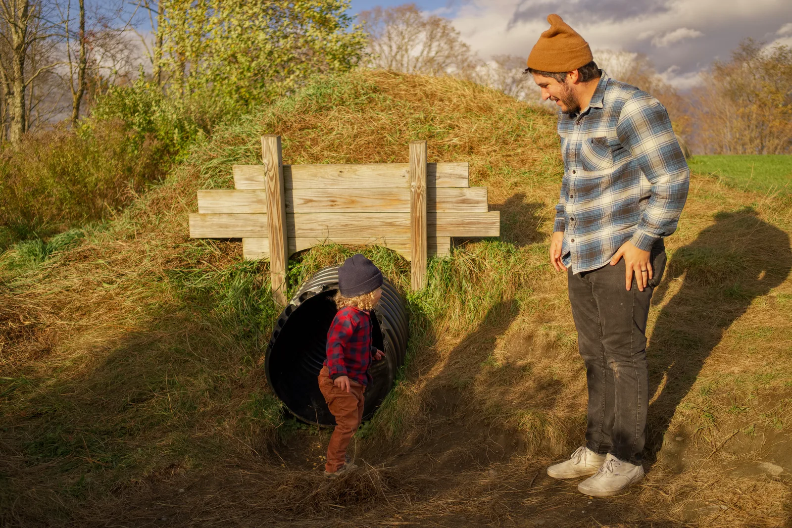 Child in red plaid shirt and brown pants climbing out of culvert, adult watching