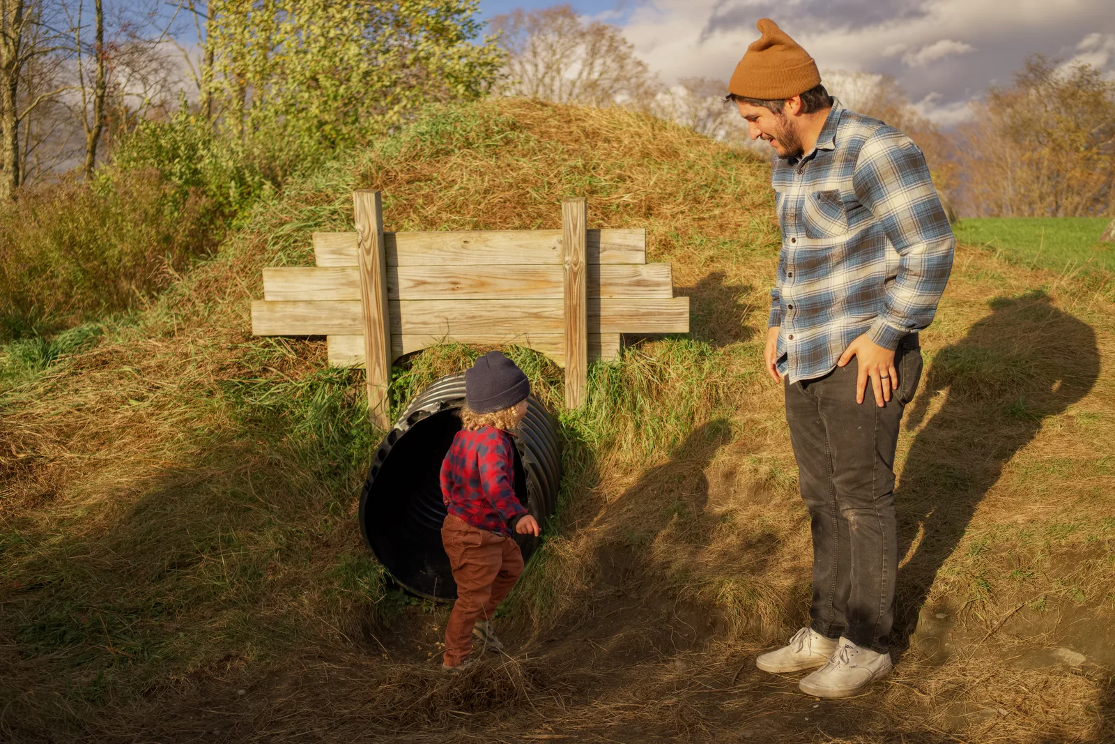 Child in red plaid shirt and brown pants at culvert entrance, adult observing