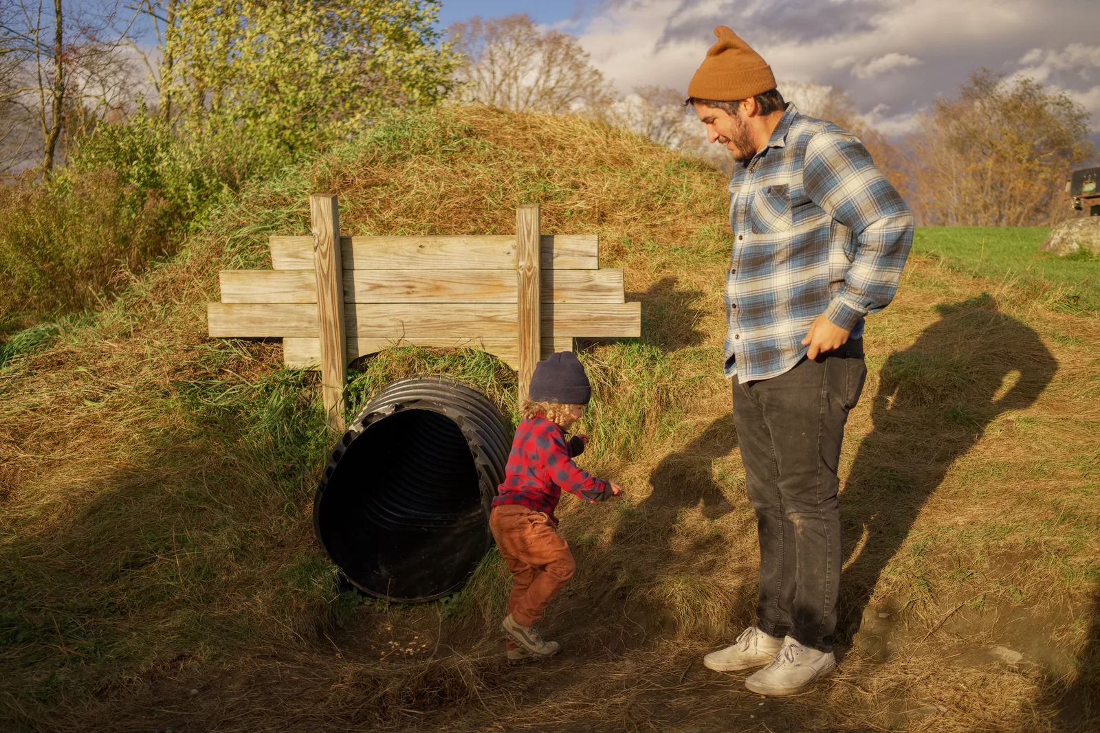 Child in red plaid shirt and brown pants near culvert, adult standing close