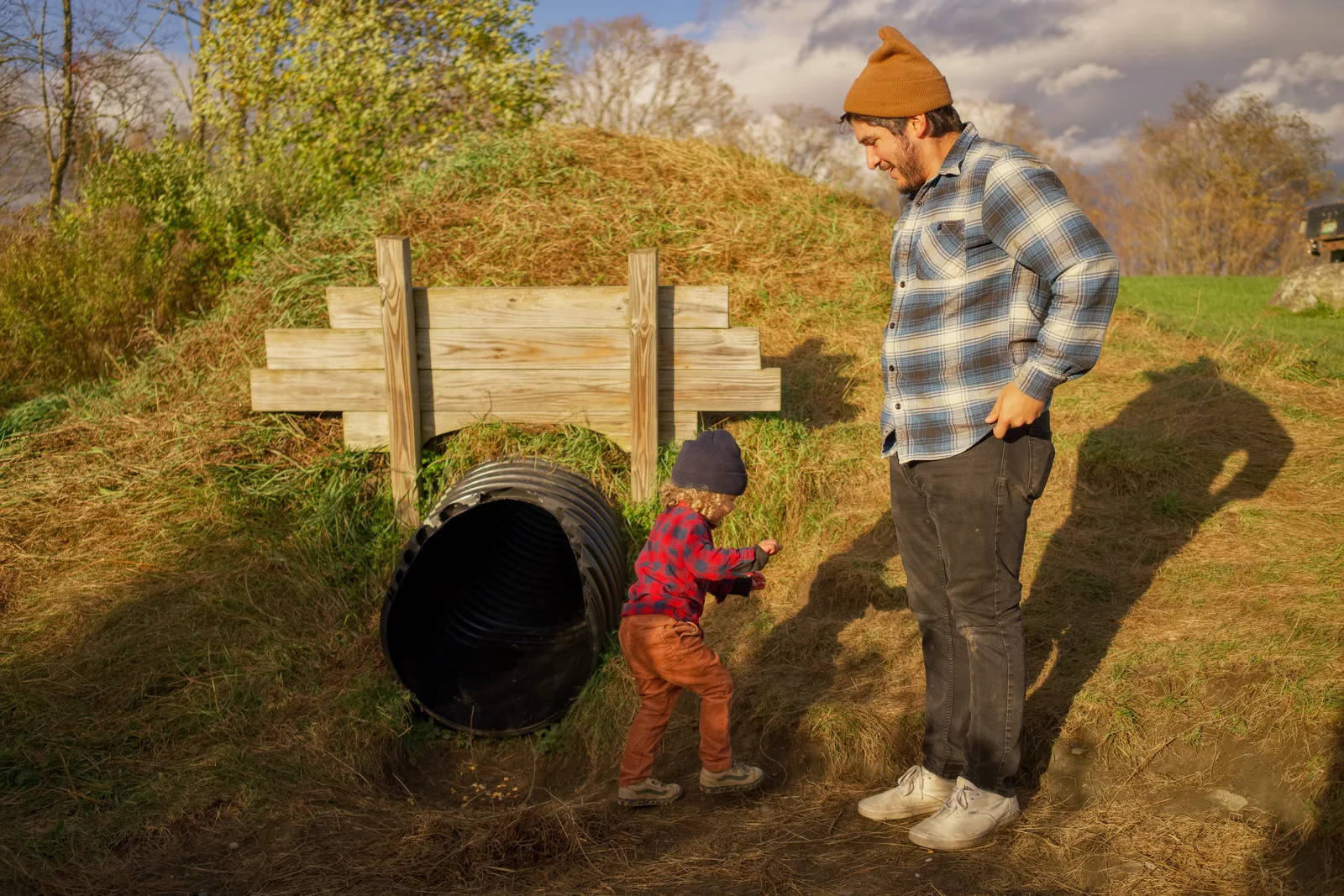 Child in red plaid shirt and brown pants pointing at culvert, adult beside