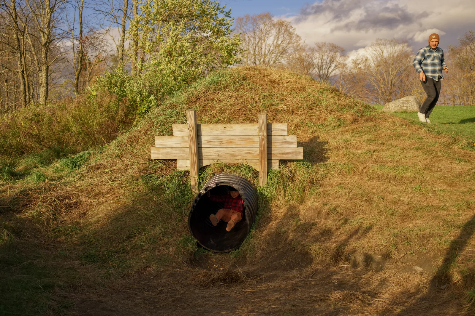 Child in red plaid shirt and brown pants lying in black culvert under wooden bridge