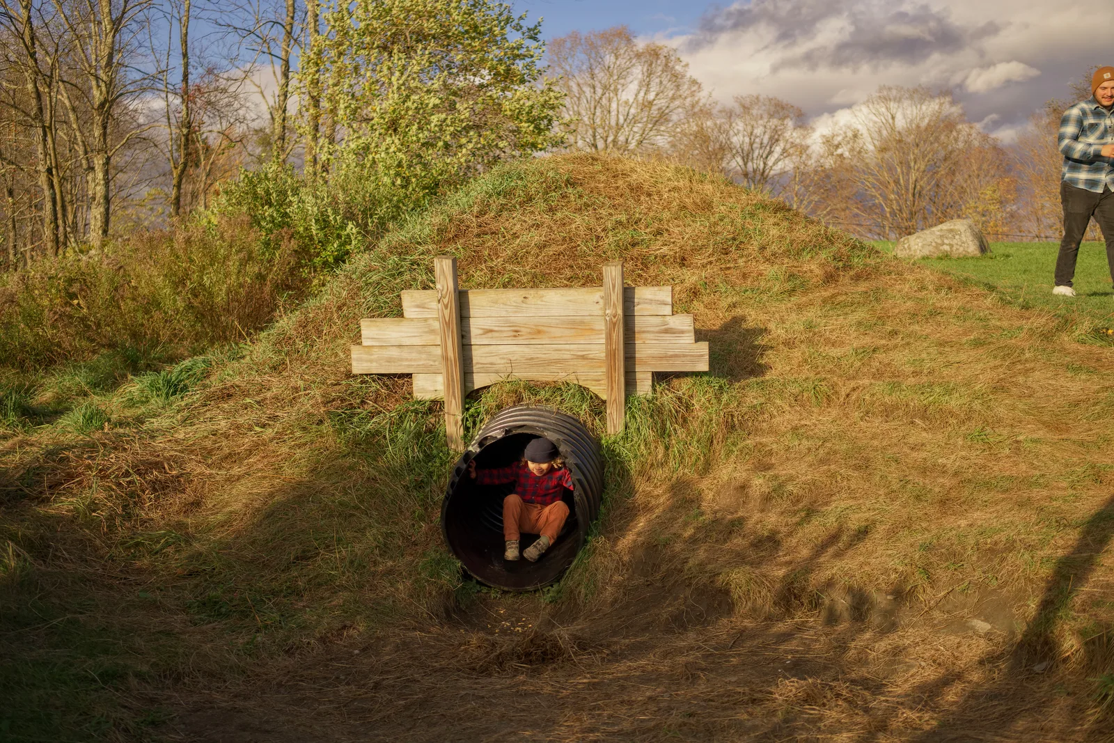 Child in red plaid shirt and brown pants sitting in black culvert under wooden bridge