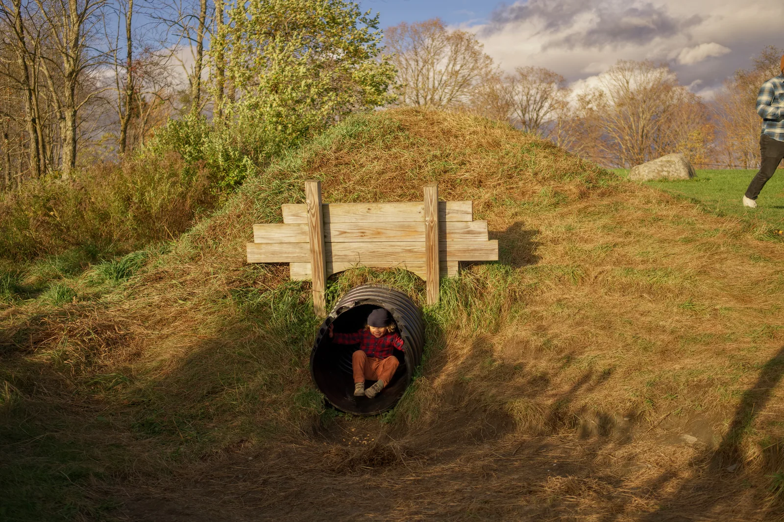 Child in red plaid shirt and brown pants crouching in black culvert under wooden bridge