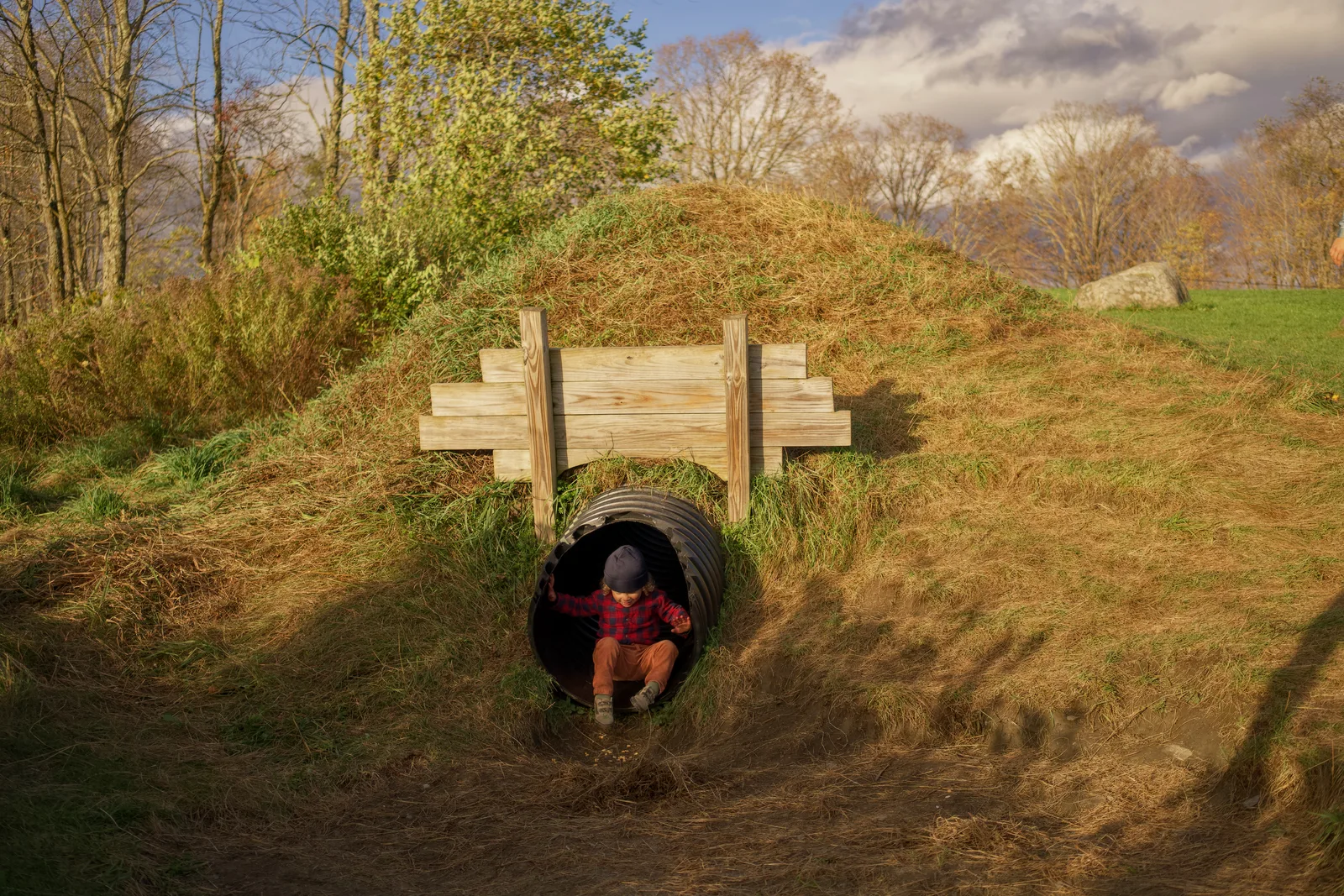 Child in red plaid shirt and brown pants crawling through black culvert under wooden bridge
