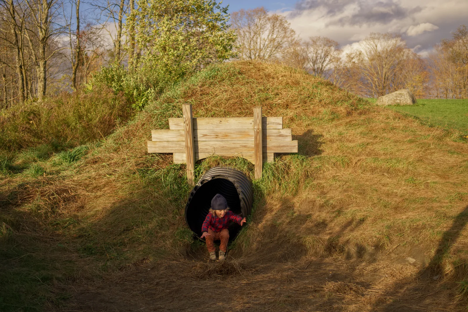 Child in red plaid shirt and brown pants exiting black culvert under wooden bridge