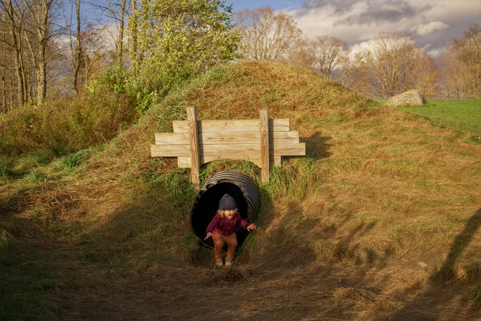 Child in red plaid shirt and brown pants walking from black culvert under wooden bridge