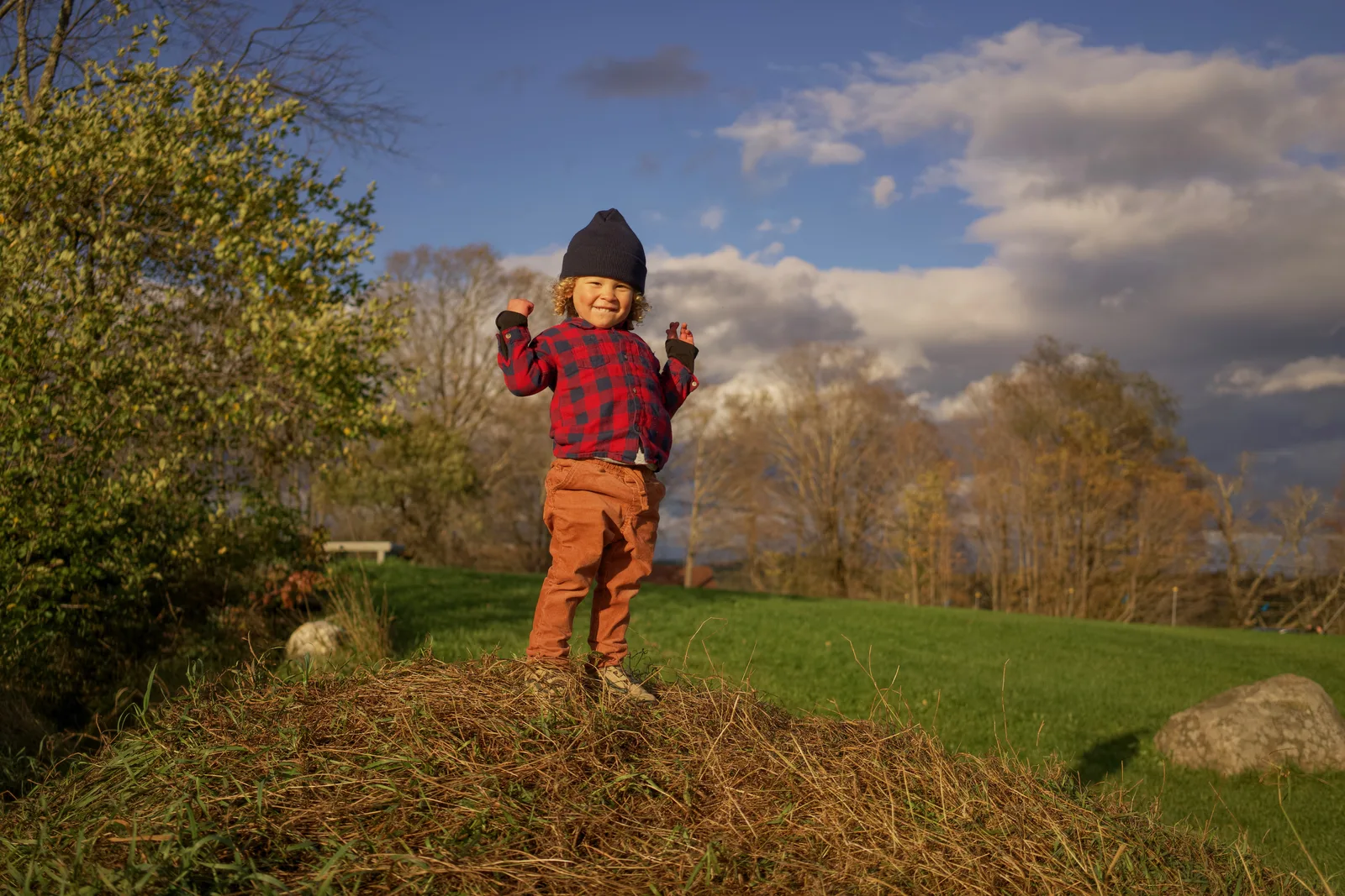 Child in red plaid shirt, brown pants, and black beanie standing on grassy hill