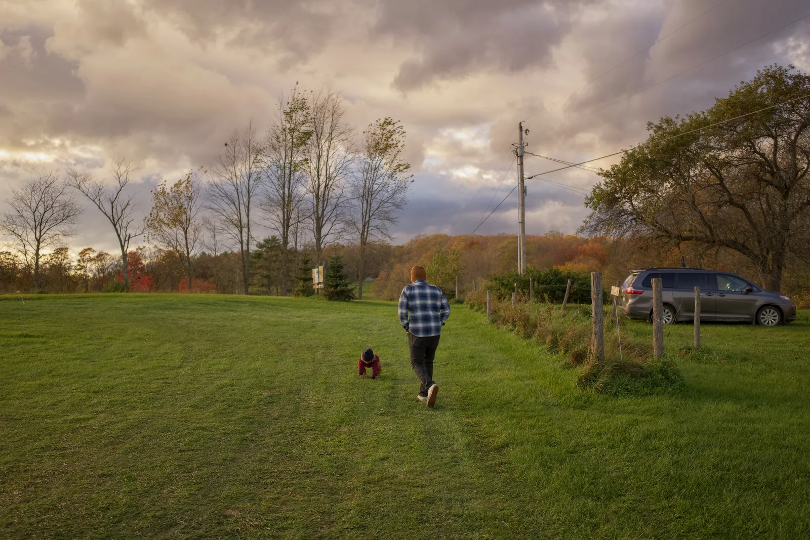 Two people walking across grassy field with car and trees under cloudy sky