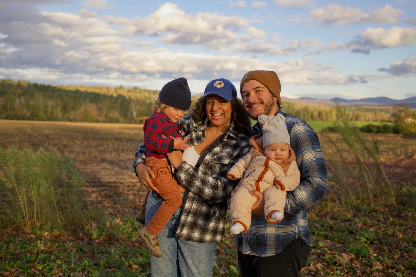 Four people in plaid and beanies holding two children in autumn field with mountains and clouds