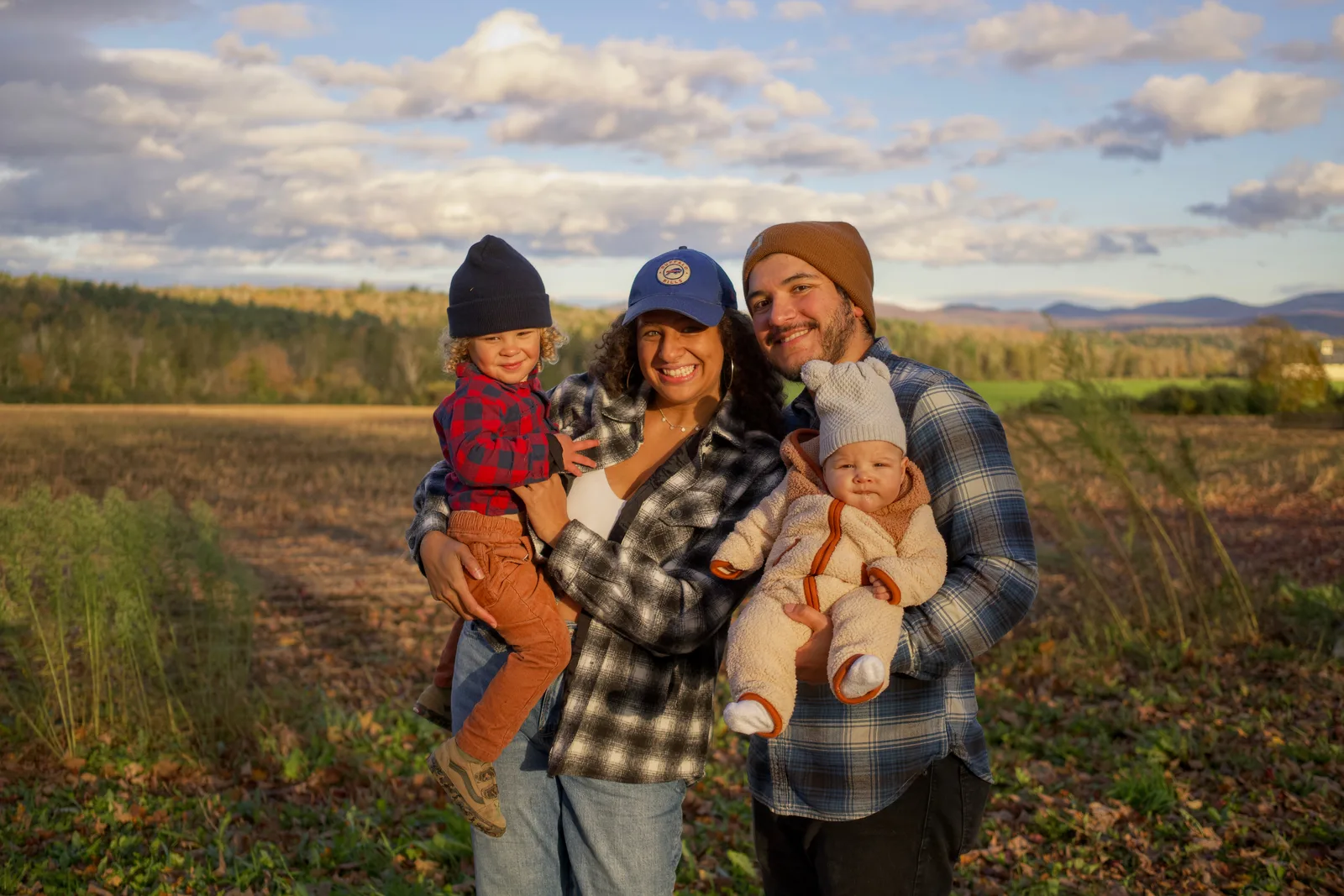 Four people in plaid and beanies holding two children in field with mountains and clouds