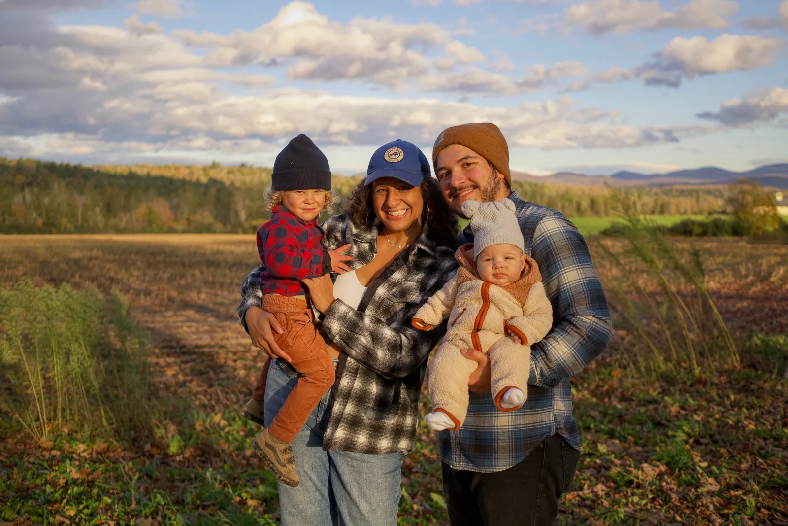 Four people in plaid and beanies holding two children in field with mountains and scattered clouds