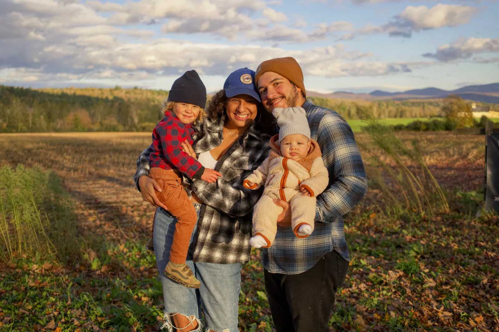 Four people in a field, two adults holding two children, forested hills in distance
