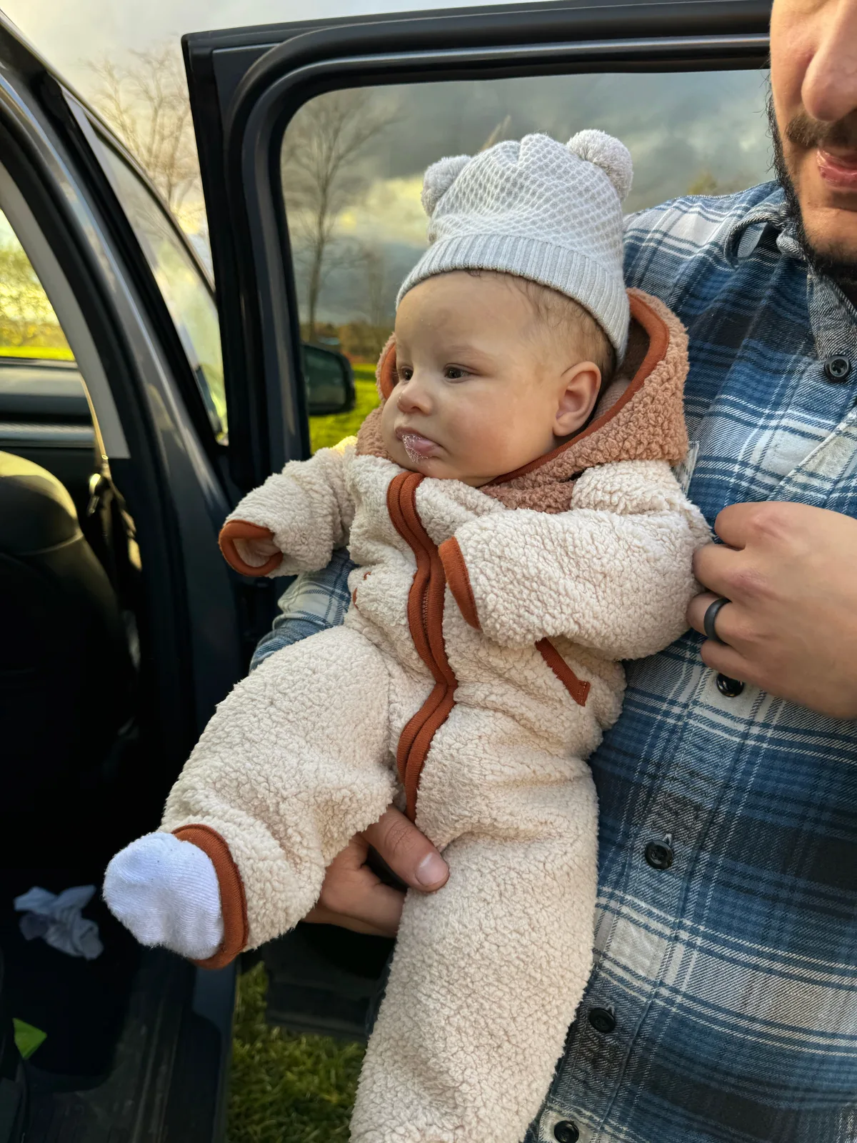 Adult in plaid shirt holding baby in white bear hat and cream onesie by open car door