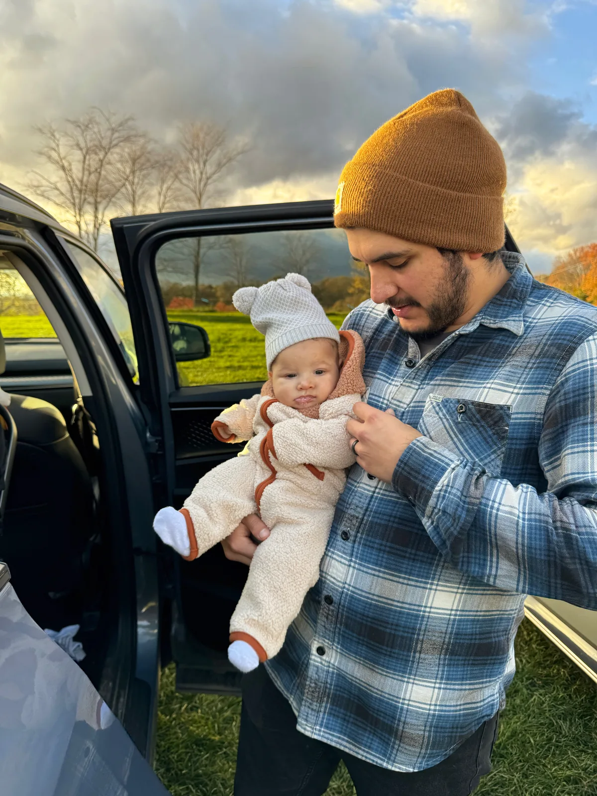 Adult in plaid shirt and brown beanie holding baby in white bear hat by car