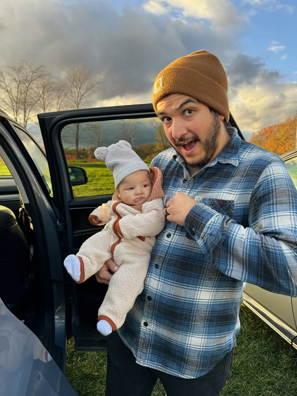 Adult in plaid shirt and brown beanie holding baby in white bear hat near open car