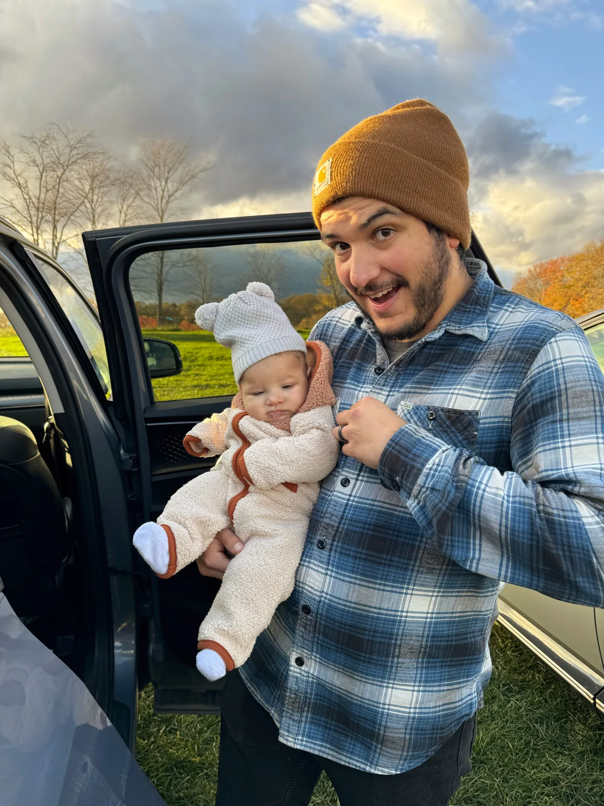 Adult in plaid shirt and brown beanie holding baby in white bear hat by car door