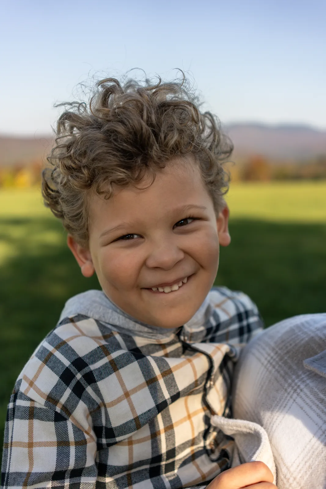 Child with curly hair smiling, wearing a plaid shirt, green field and mountains behind