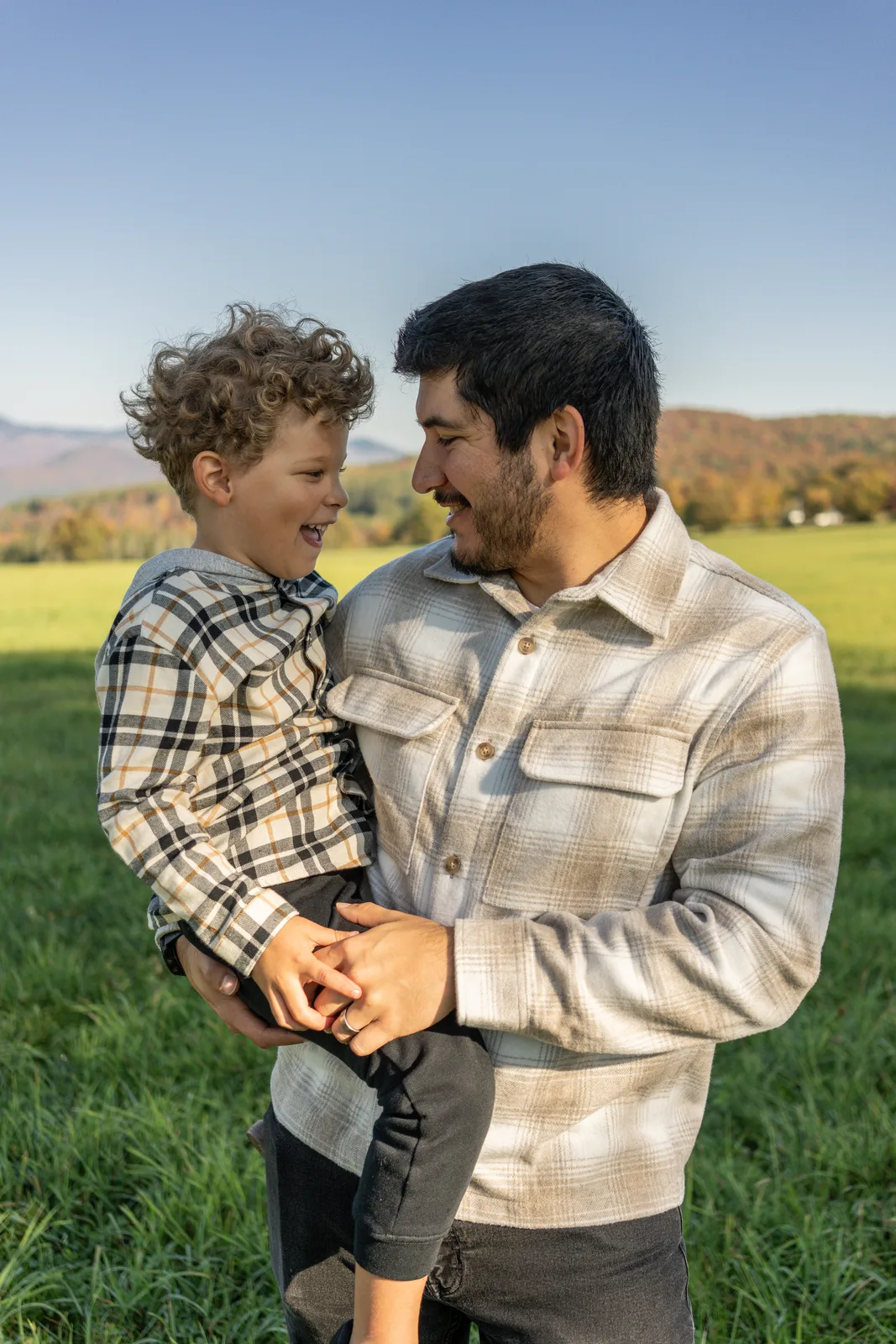 Adult holding a child with curly hair, both smiling, green field and mountains behind