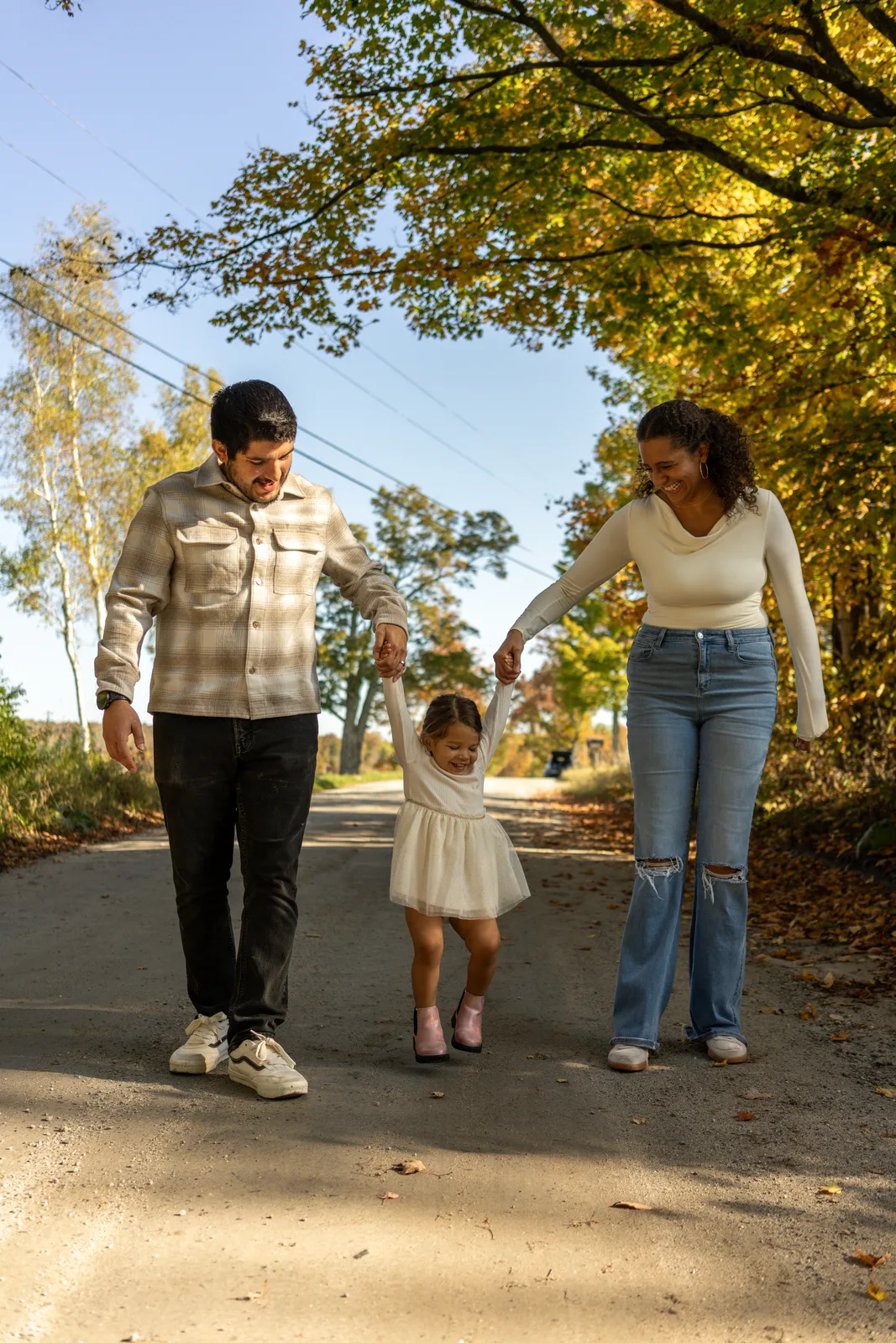 Three people walking on a sunlit road, holding hands with a child in a white dress between them, surrounded by autumn trees.