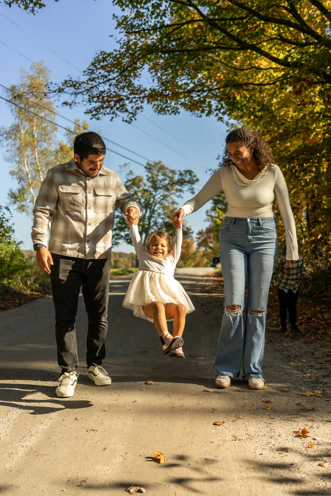Three people walking on a road, lifting a child in a white dress between them, with fall foliage overhead.