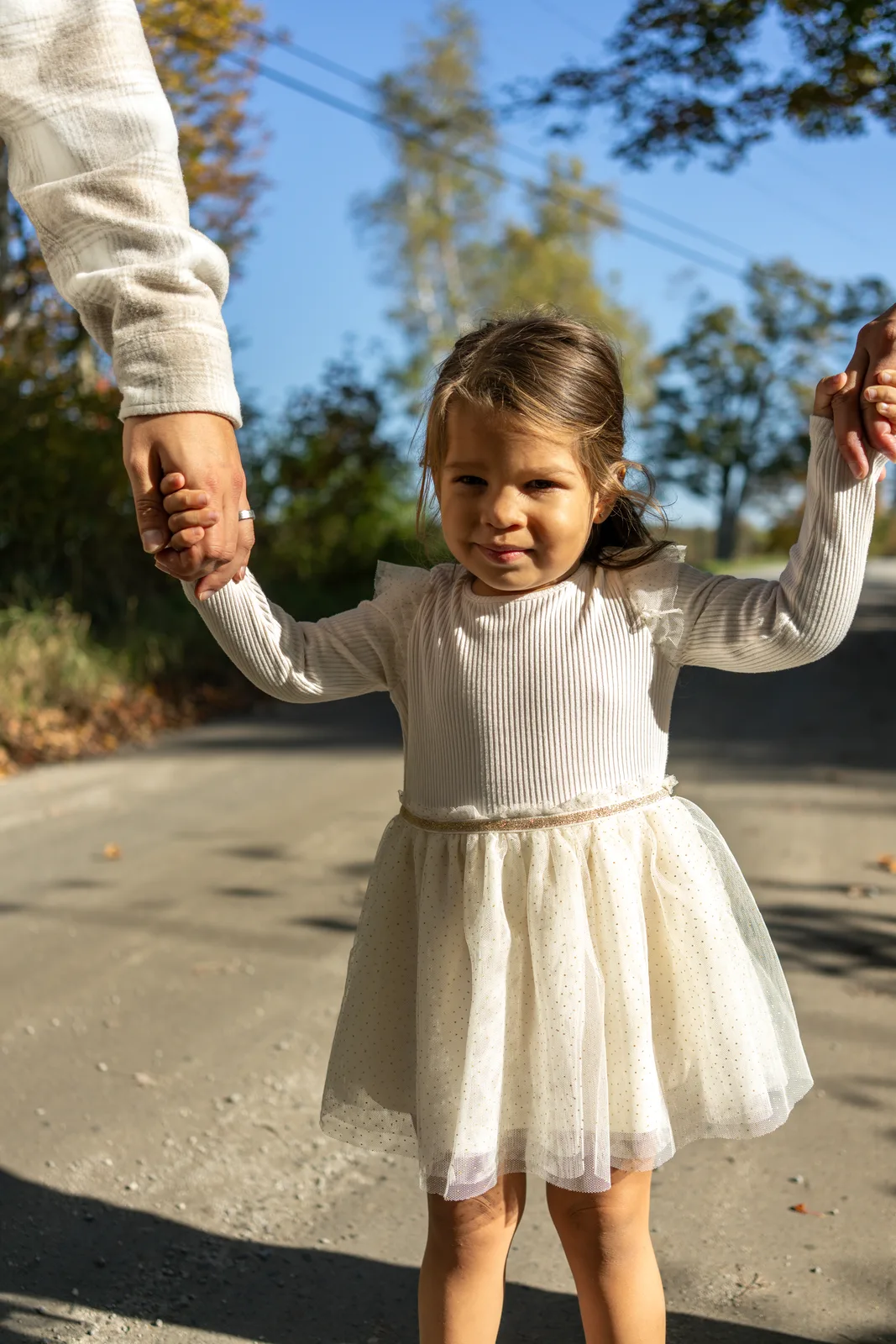 Child in a white dress and long sleeves holding hands with two adults on a sunlit road, trees in background.