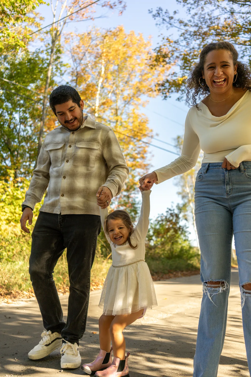 Three people walking on a road, holding hands with a child in a white dress, surrounded by autumn trees.