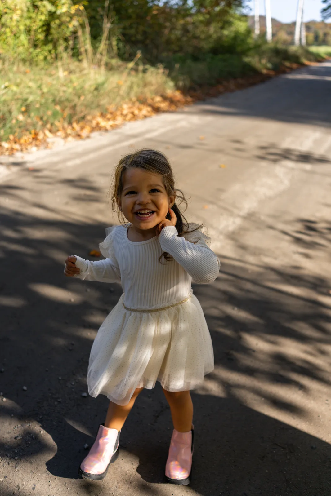 Child in a white dress and pink boots running on a sunlit road, with trees and fallen leaves around.