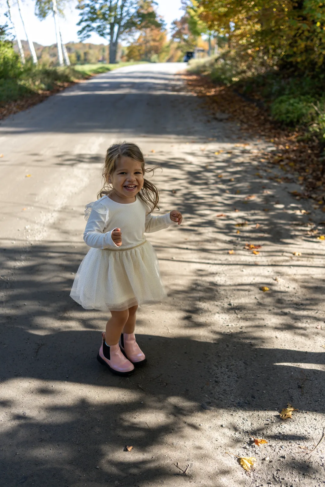 Child in a white dress and pink boots running on a sunlit road, with trees and shadows on the ground.