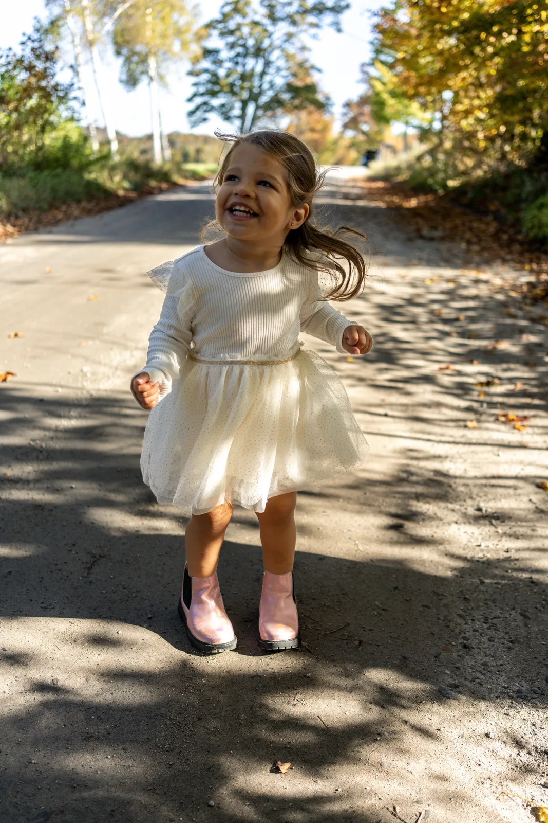 Child in a white dress and pink boots running on a sunlit road, with autumn trees in the background.