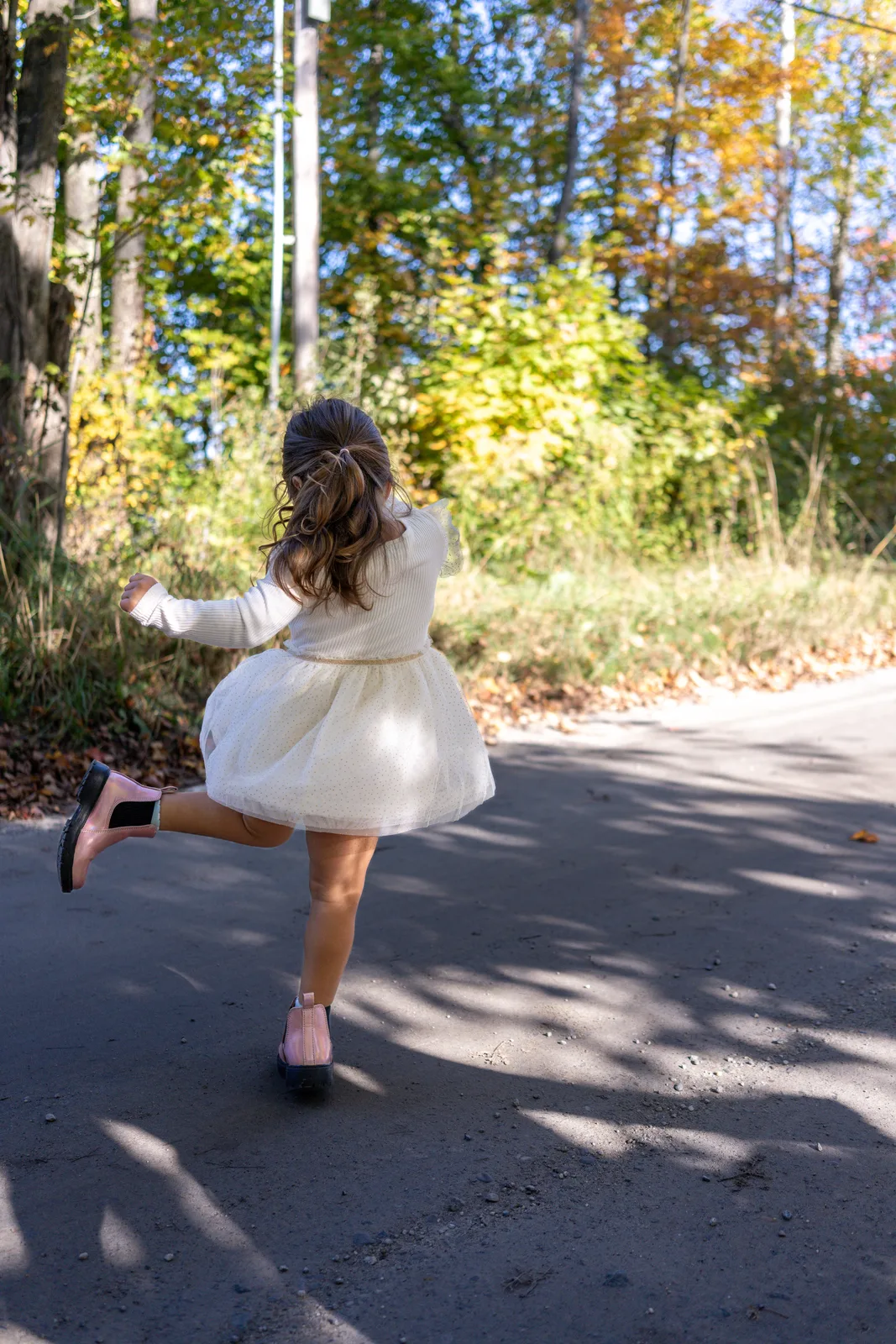 Child in a white dress and pink boots running away on a sunlit road, with trees and shadows around.