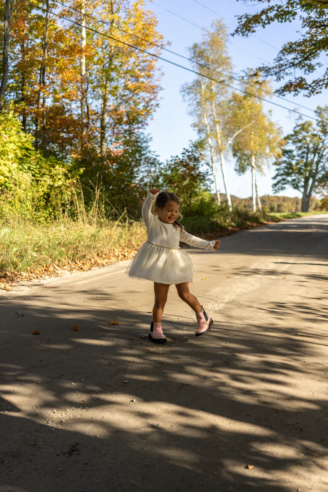 Child in white dress and pink shoes twirling on a sunlit road with autumn trees