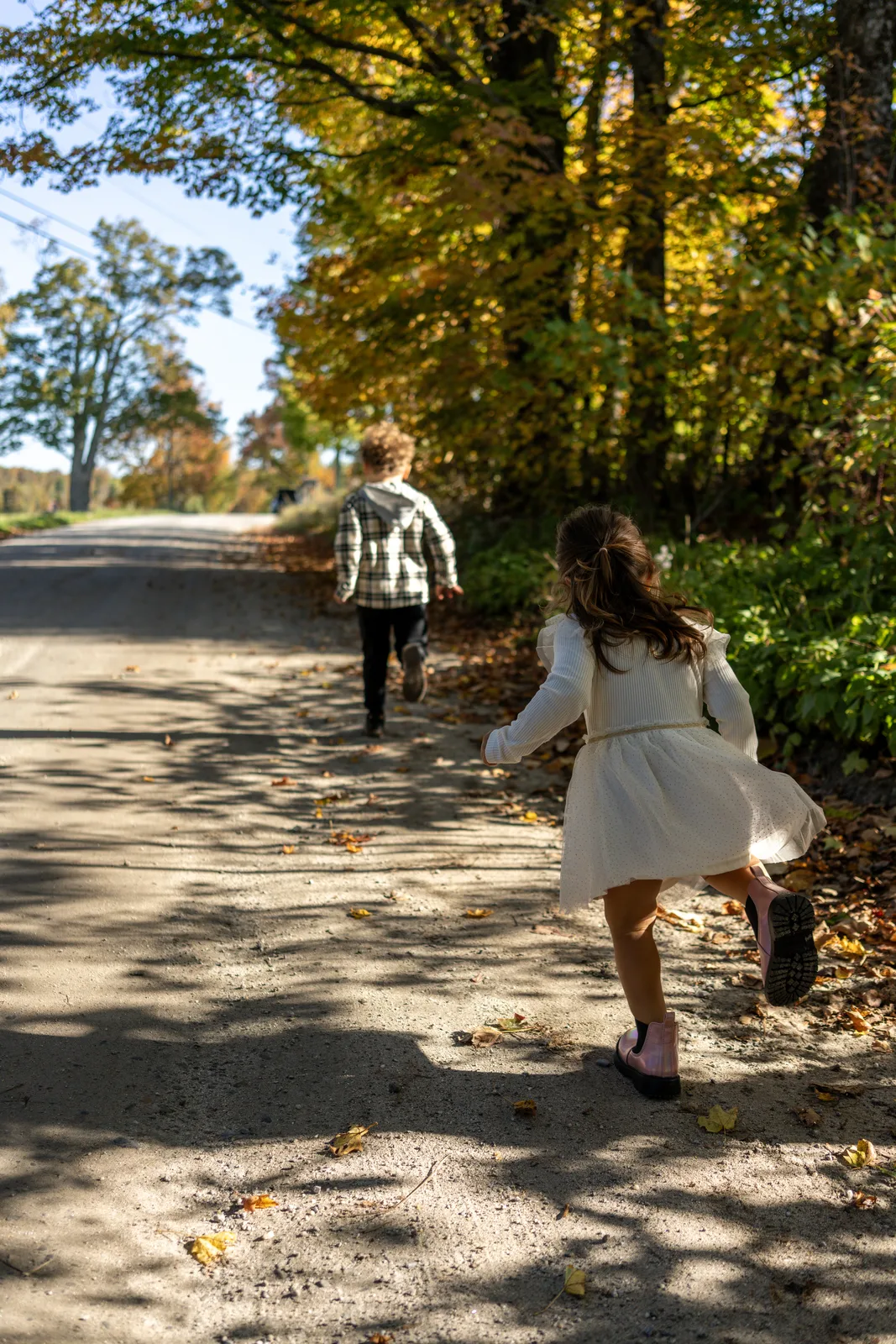 Two children running on a shaded road, one in a white dress, one in plaid shirt