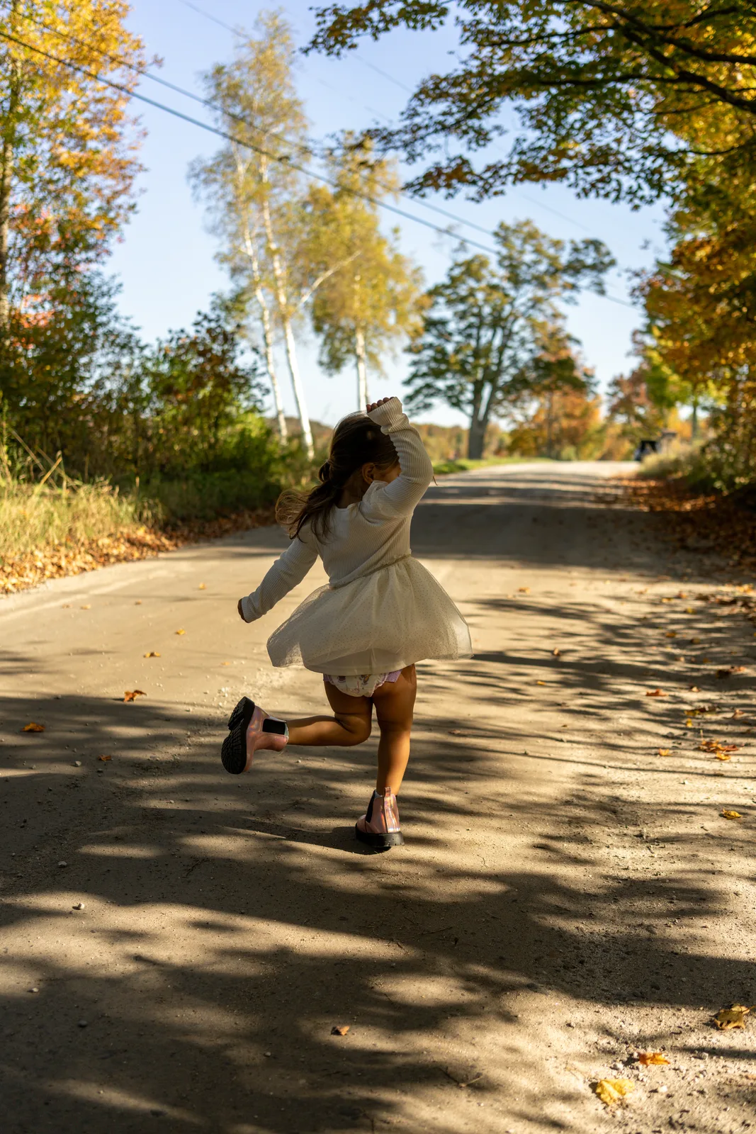 Child in white dress and black shoes running on a sun-dappled road
