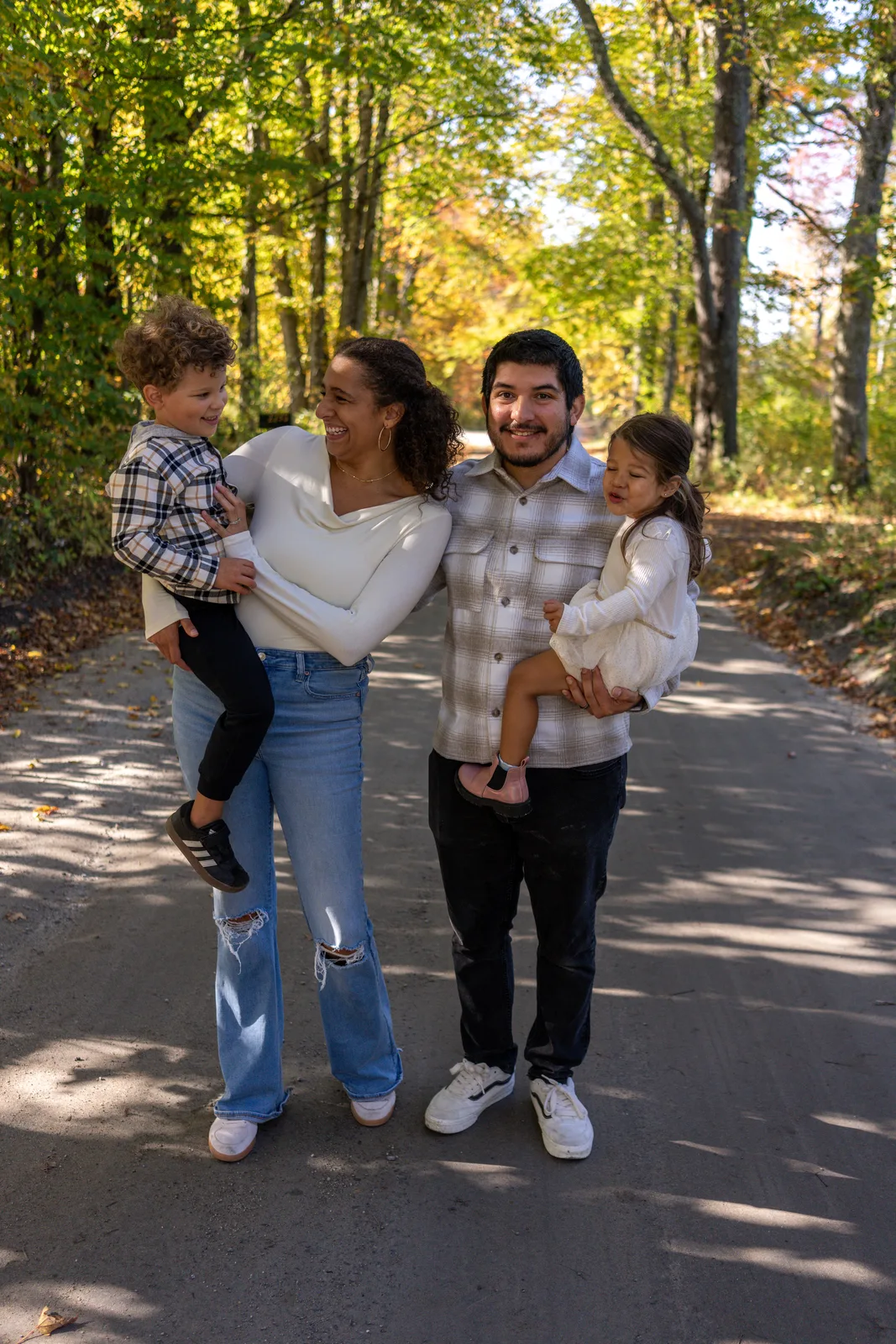 Four people on a sunlit wooded road, two adults holding two children in plaid and white