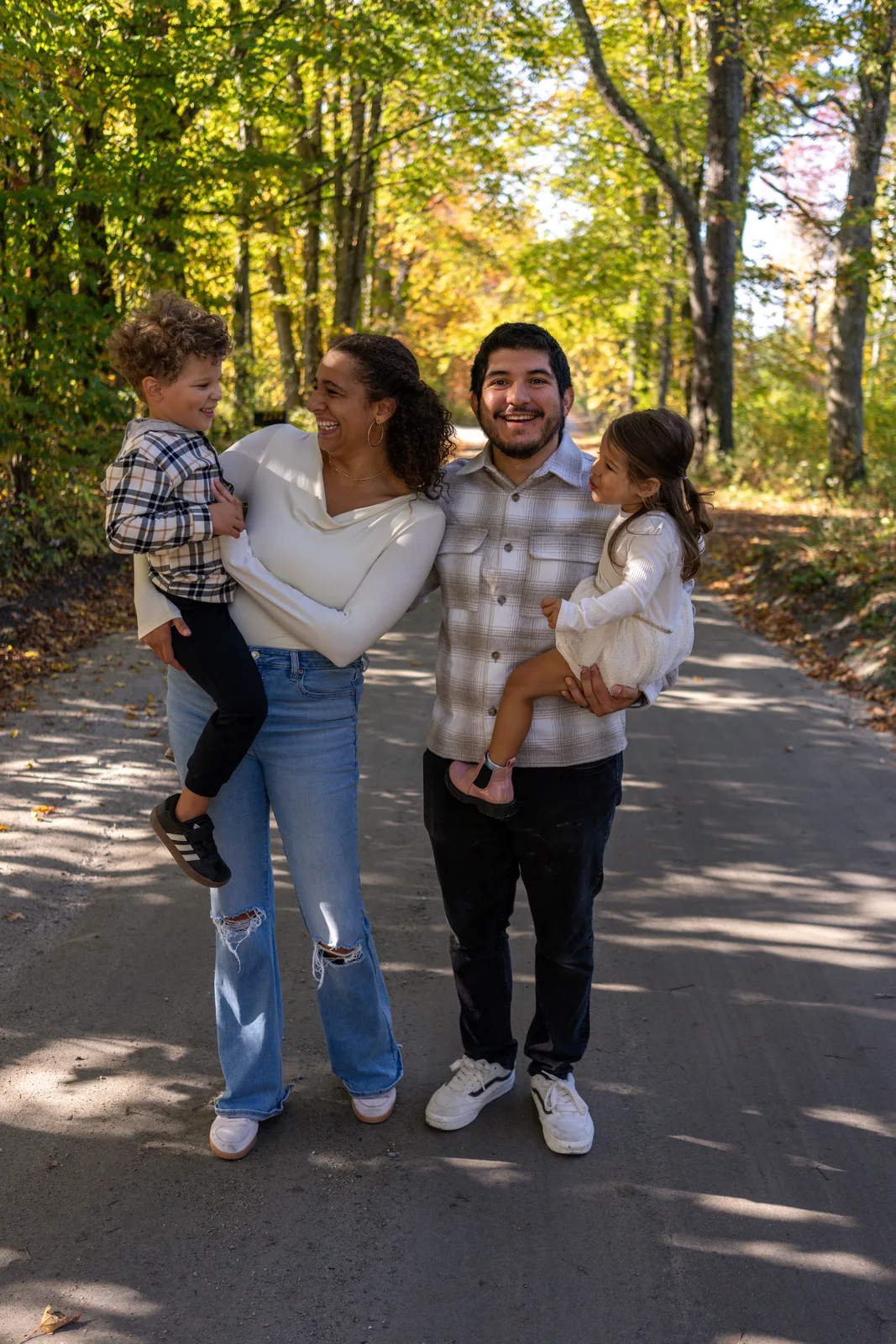 Four people on a shaded wooded road, two adults holding two children in plaid and white