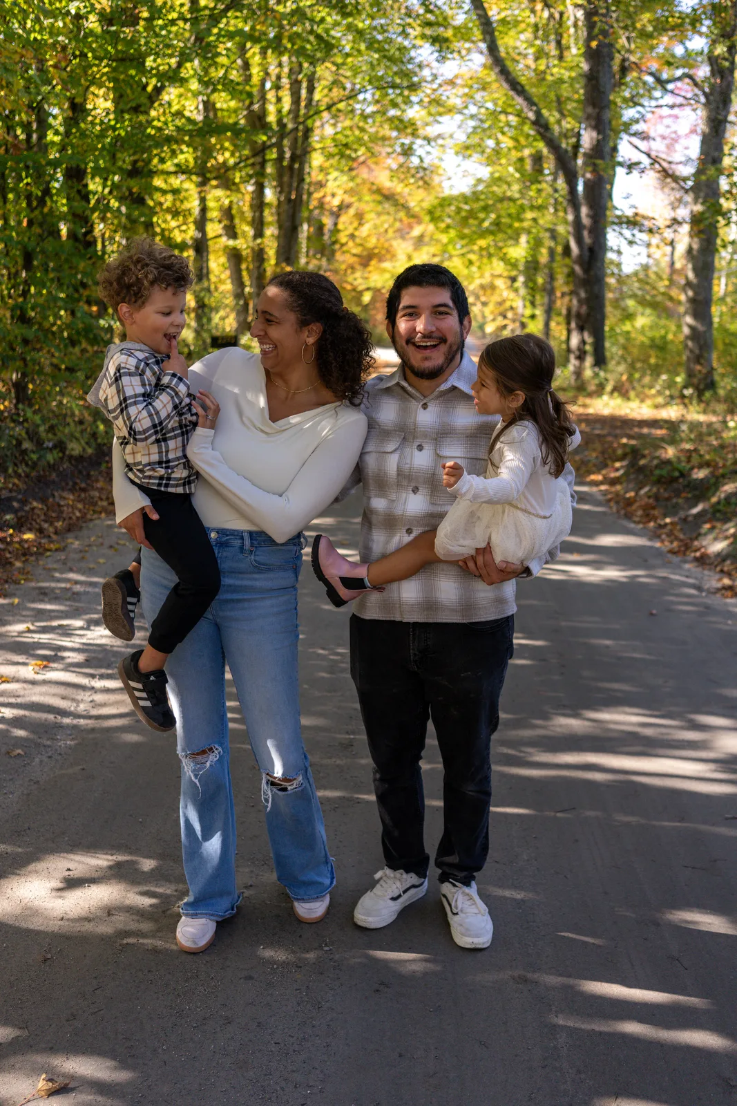Four people on a dappled wooded road, two adults holding two children in plaid and white