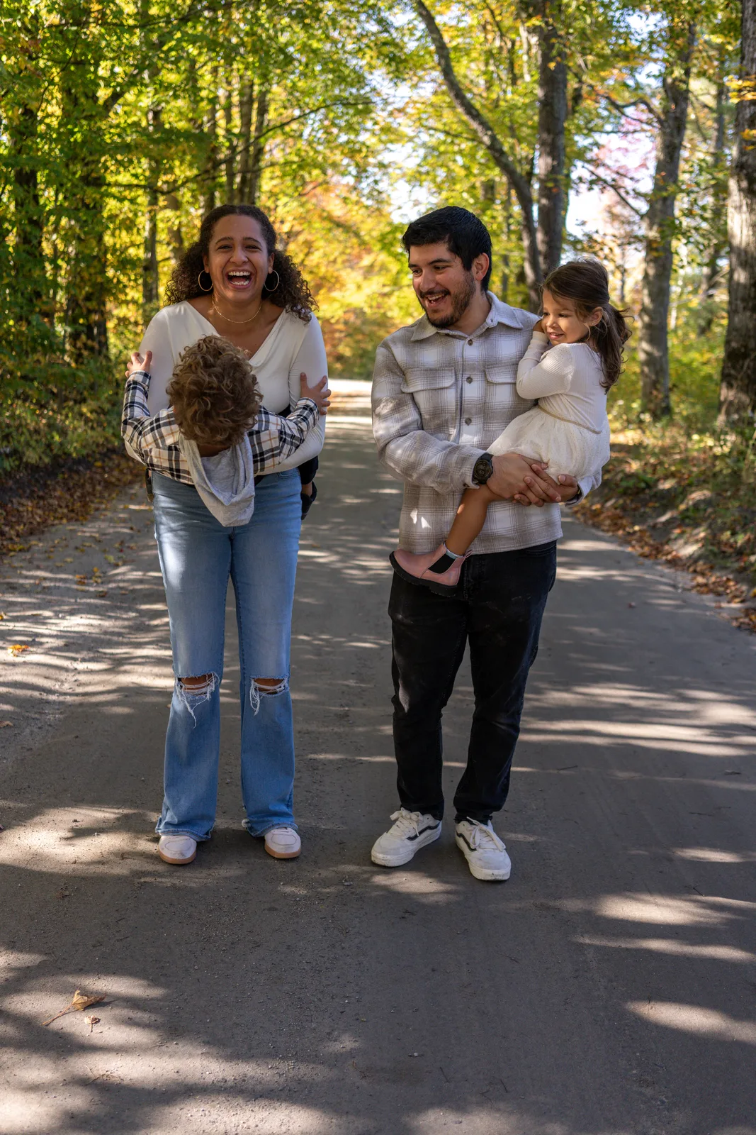 Two adults walking on a wooded road, each holding a child in plaid and white