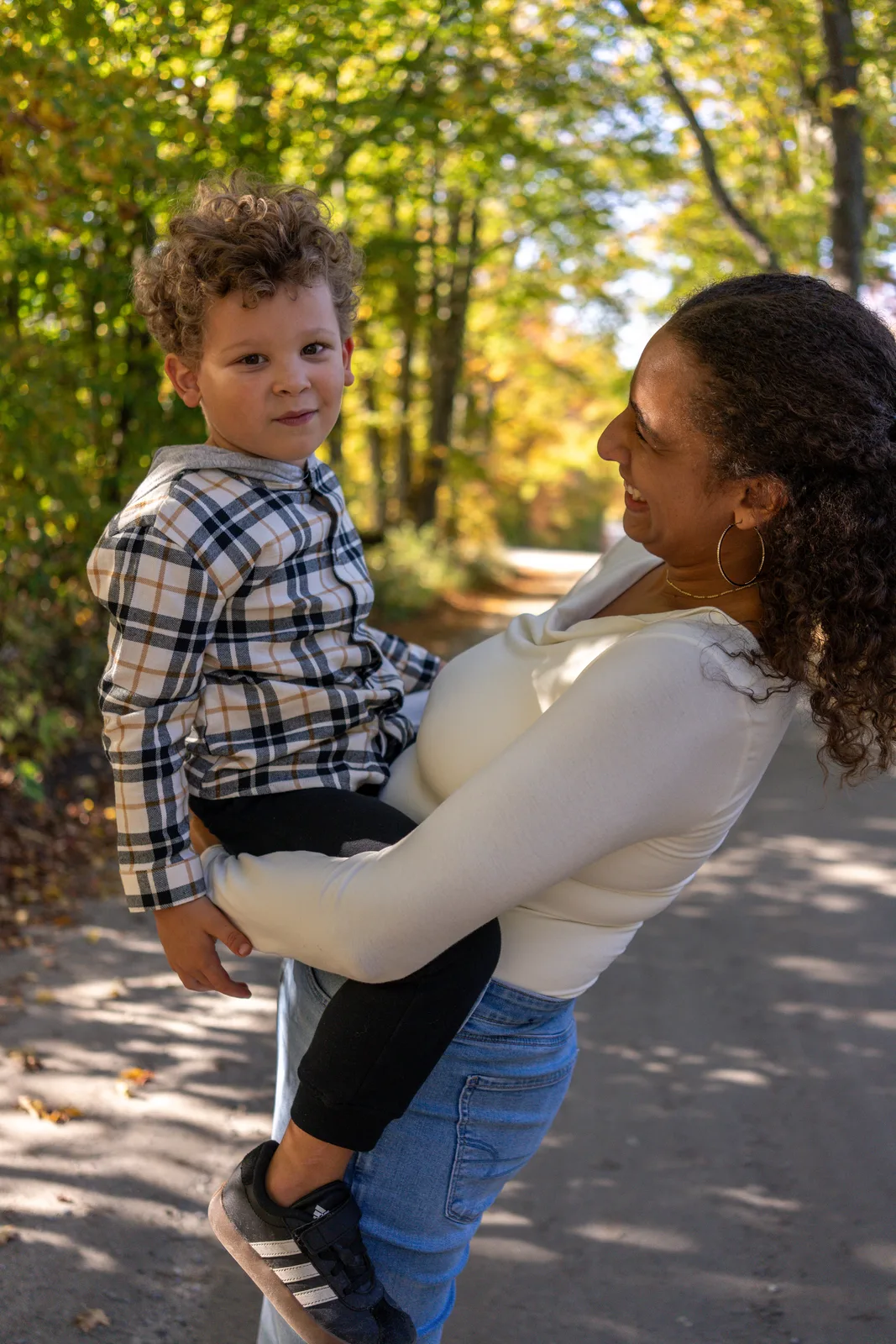 Adult in white top and blue jeans holding a child in plaid shirt on a wooded path