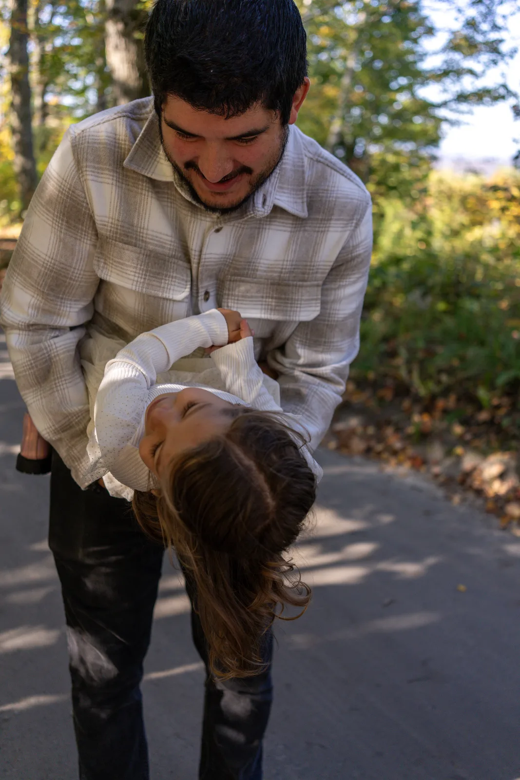 Adult in plaid shirt holding a child upside down on a sunlit wooded path