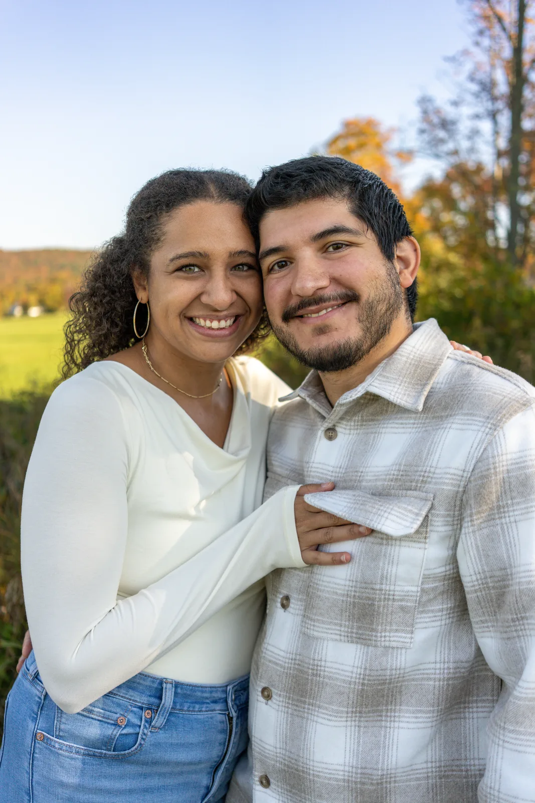 Two adults embracing in a field with autumn trees and mountains in the background